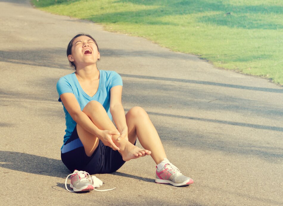 Young woman sits on ground with running shoe off, holding sprained ankle 