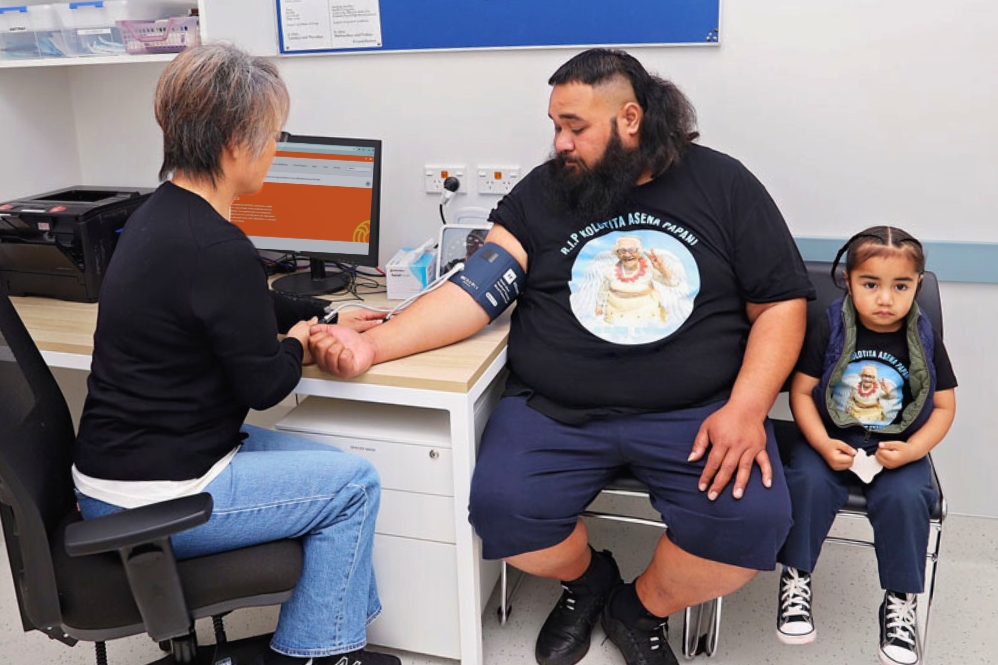 Healthcare provider taking blood pressure of a Pasifika male accompanied by young son, in a clinic setting.