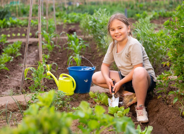 Girl crouched down in home vegetable garden with bucket, watering can and trowel