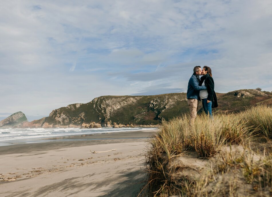 Pregnant couple kissing on grassy dunes by beach